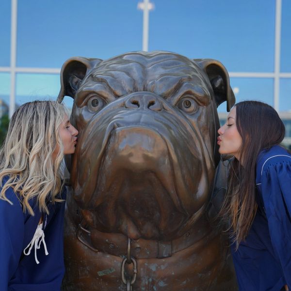 Two women in blue gowns kiss a large bronze bulldog statue outdoors.