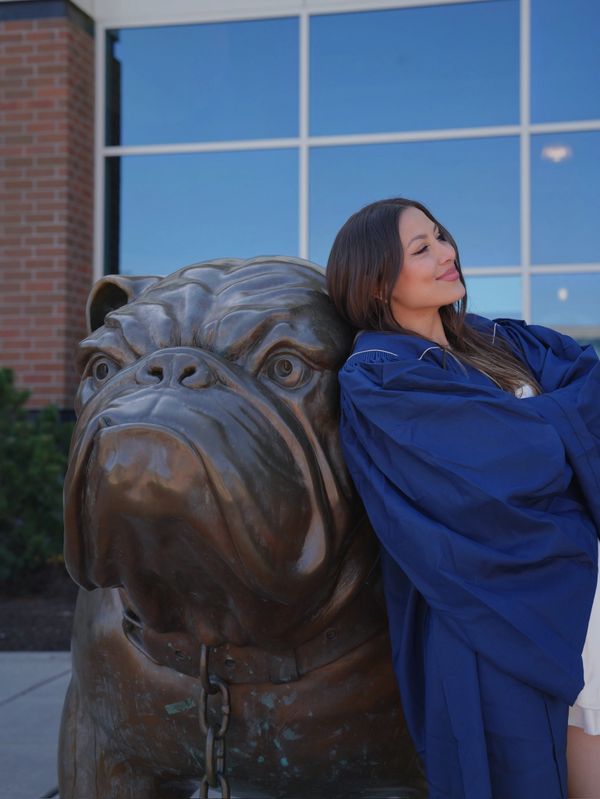 A woman in a blue graduation gown leans against a large bulldog statue.