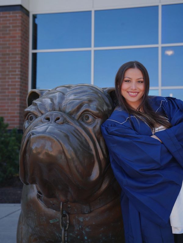 A graduate in a blue gown poses cheerfully beside a large bulldog statue.