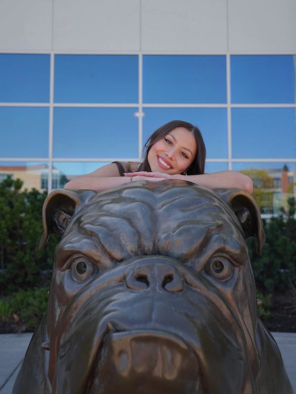 A smiling woman leans on a large bulldog statue outdoors.