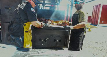 Two men carrying a large tray of cooked meat outdoors.