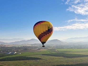 A colorful hot air balloon floats above green fields with mountains in the background under a clear sky.
