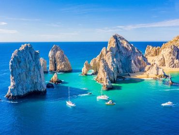 Sailboats near rocky cliffs in clear blue ocean waters under a sunny sky.