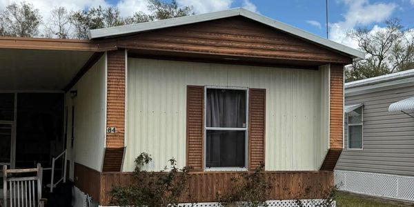 A beige and brown mobile home with a single window and small shrubs in front.