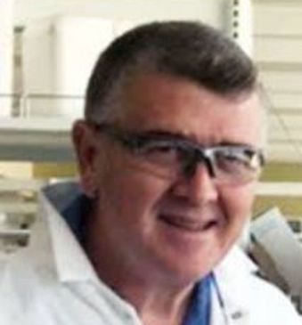 A man wearing safety glasses and a lab coat smiles in a laboratory.