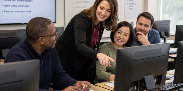 A teacher assists three adult students at a computer in a classroom.
