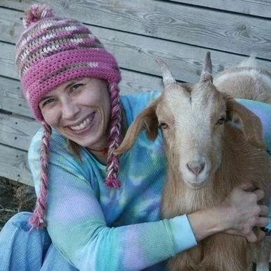 Smiling woman in a pink knit hat hugs a brown goat.