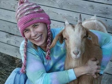 Smiling woman in a pink knit hat hugs a brown goat.