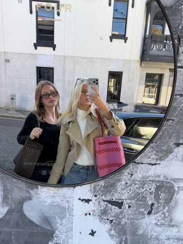 Two young women taking a mirror selfie on the street with stylish bags.