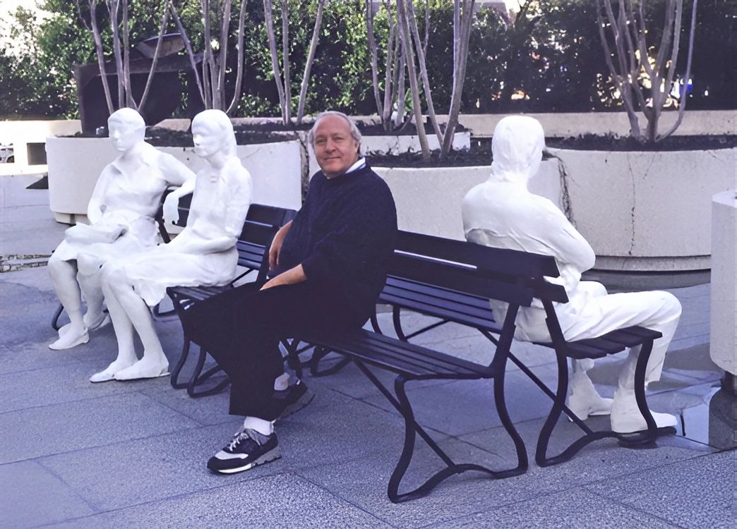 Man sits on a bench with three white human-like sculptures, two seated and one with arms crossed.
