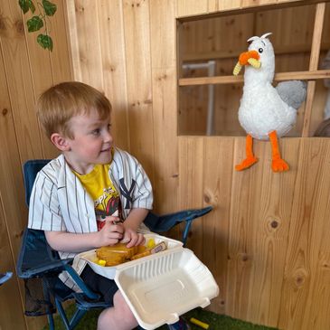 Young ginger boy eating pretend fish and chips on a camping chair with a toy seagull watching him.
