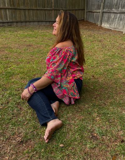 Woman sitting barefoot on grass wearing a colorful top and jeans.