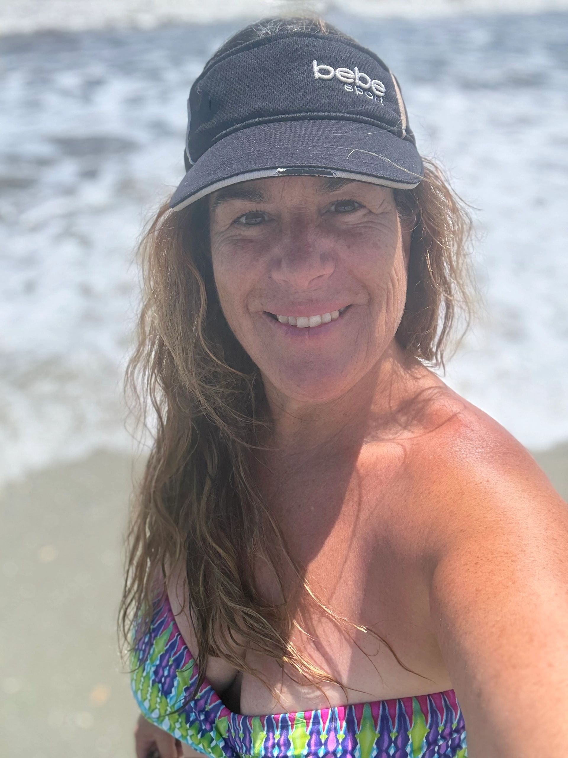 Woman smiling at the beach wearing a sun visor and colorful swimsuit.