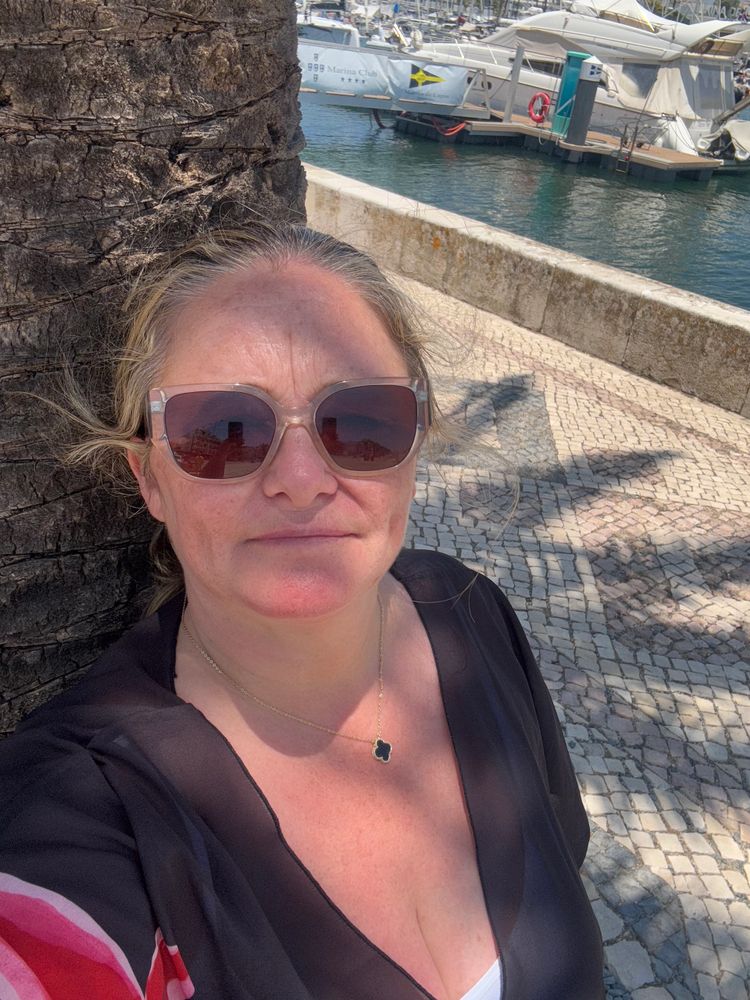 Woman wearing sunglasses leans against a tree by a marina on a sunny day.