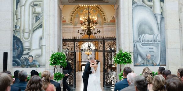 A couple shares a kiss during their wedding ceremony in an ornate indoor venue.