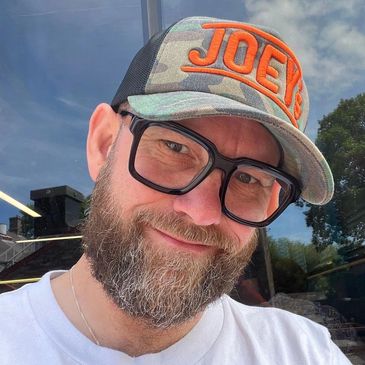 Smiling man with beard and large glasses wearing a camo hat.