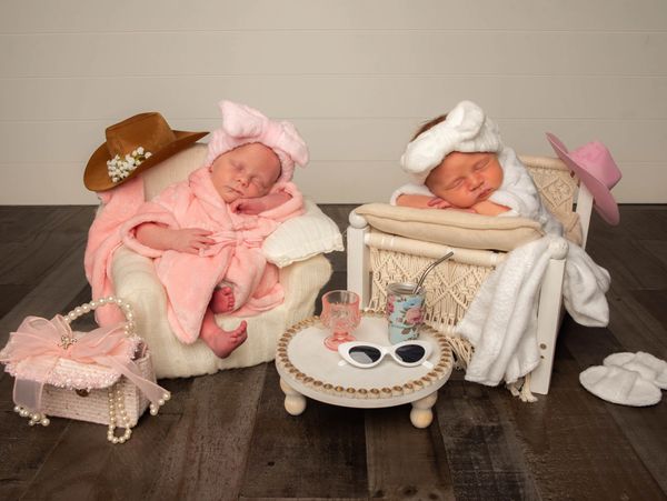 Two newborns dressed in robes and headbands, peacefully sleeping in miniature chairs.