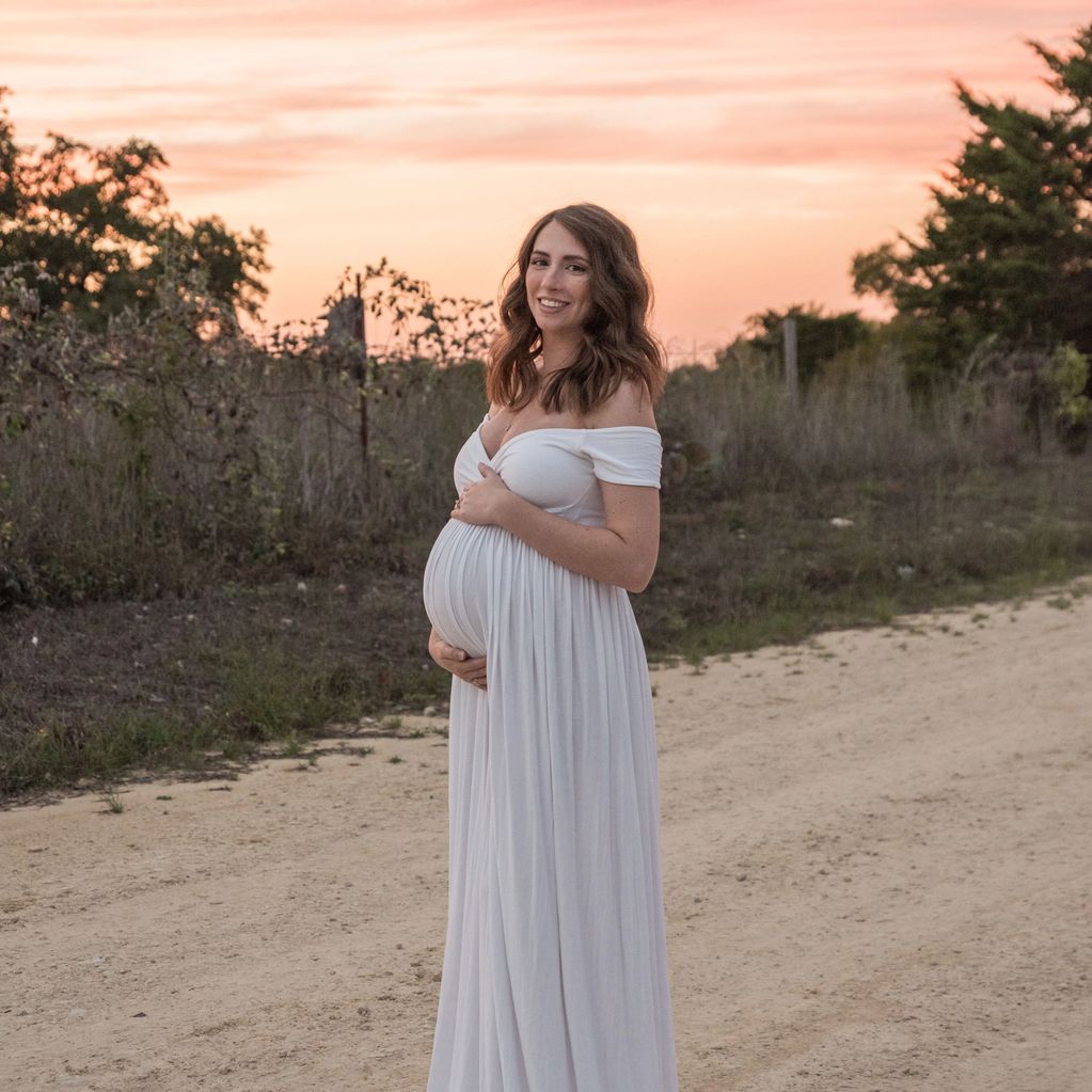 Pregnant woman in a white dress smiles at sunset on a country path.