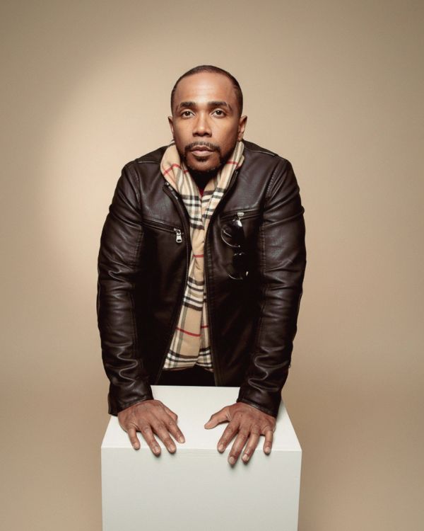 Man in black leather jacket leaning on a white pedestal against a beige background.