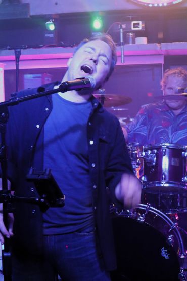 Man passionately singing with drummer in the background under colorful stage lights.
