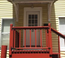 A red wooden porch with railing and stairs in front of a yellow house.