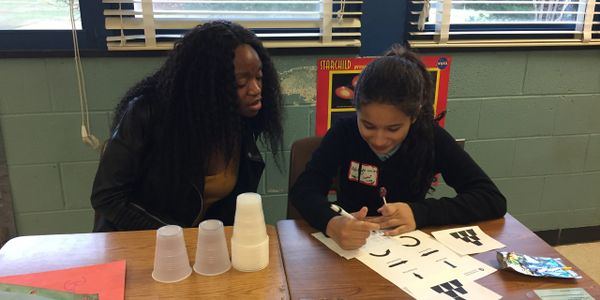 A woman watches a girl drawing symbols on paper at a table.