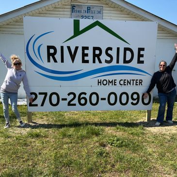 Two women stand on either side of a Riverside Home Center sign outdoors.