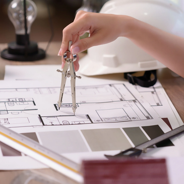 Hand using a compass on architectural blueprints with a hard hat nearby.