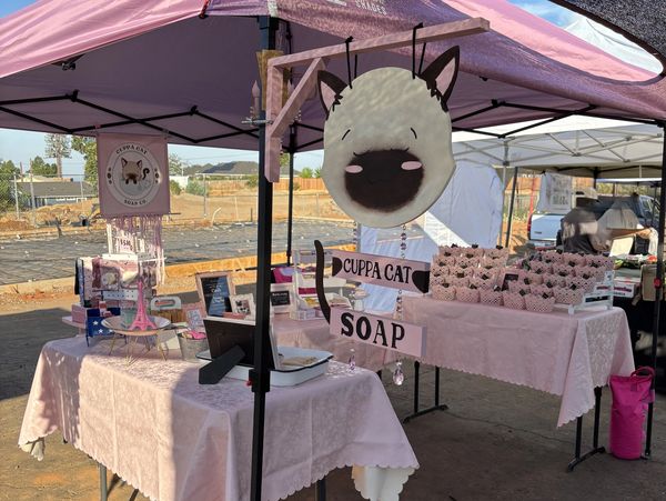 Outdoor market stall selling Cuppa Cat soap with pink decorations.