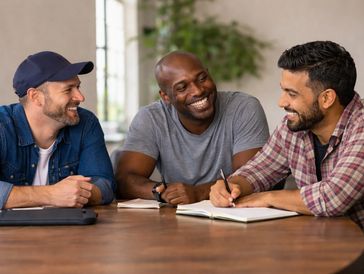 Three men smiling and chatting while writing at a table.