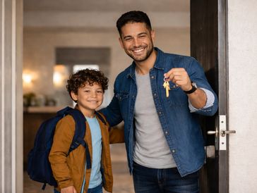 Smiling man holding house keys with a happy boy wearing a backpack at the doorway.