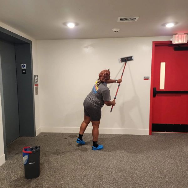 A woman cleaning a wall near a red exit door and an elevator.