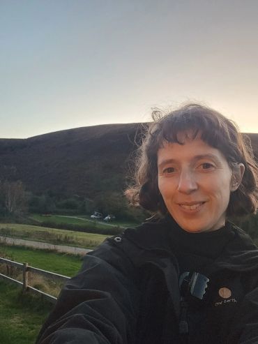 Woman smiling outdoors with hills and fields at sunset in the background.