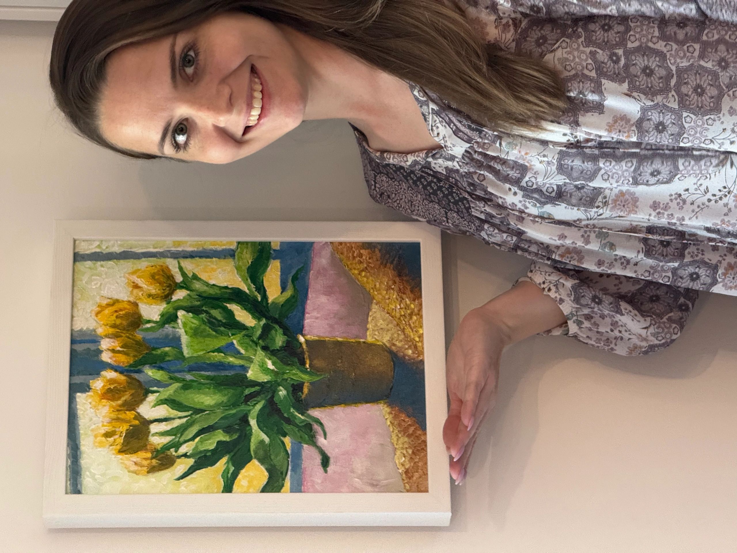 Smiling woman presenting a framed painting of yellow flowers in a vase.