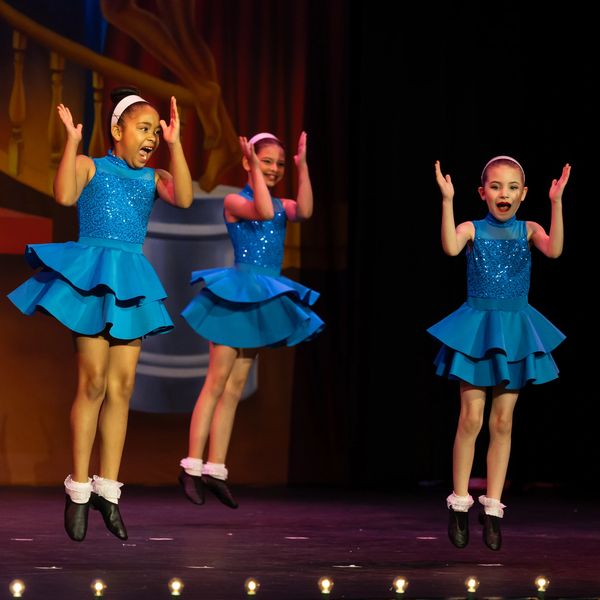 Three young girls in blue costumes performing a lively dance on stage.