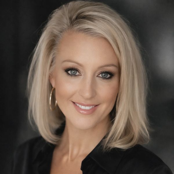 Blonde woman with blue eyes wearing a black shirt and hoop earrings smiles warmly.