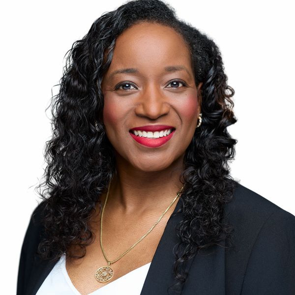 Confident woman with curly hair smiles warmly, wearing a black blazer and gold pendant necklace.