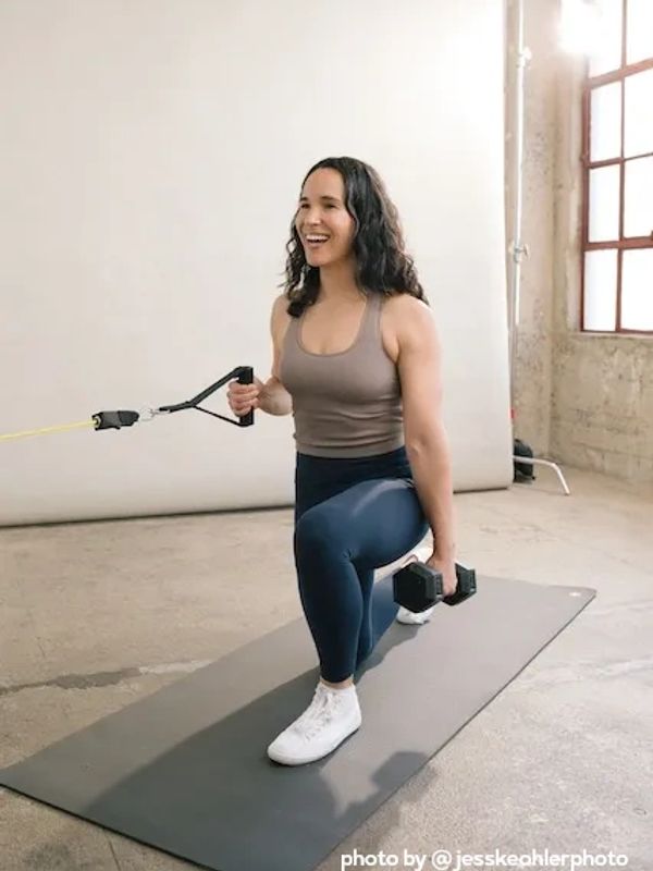 smiling mother half kneeling on a yoga mat, grasping a dumbbell and pulling resistance band. 
