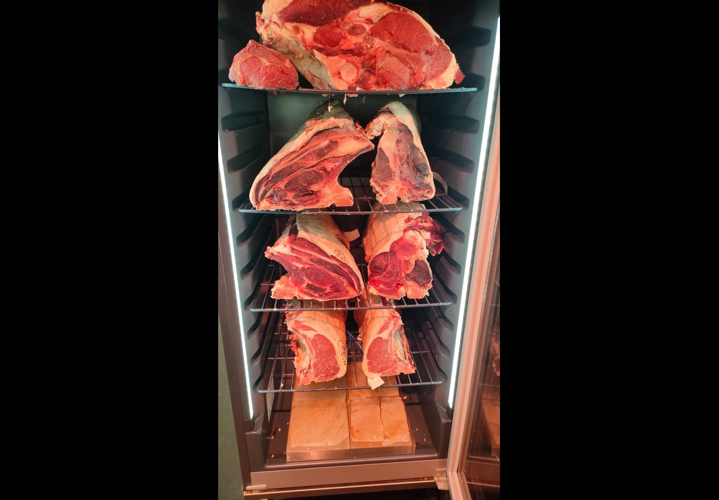 Large cuts of meat aging on racks inside a temperature-controlled fridge.
