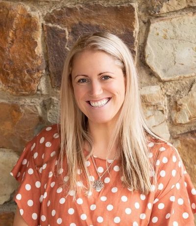 Smiling woman in a polka dot top stands against a stone wall.