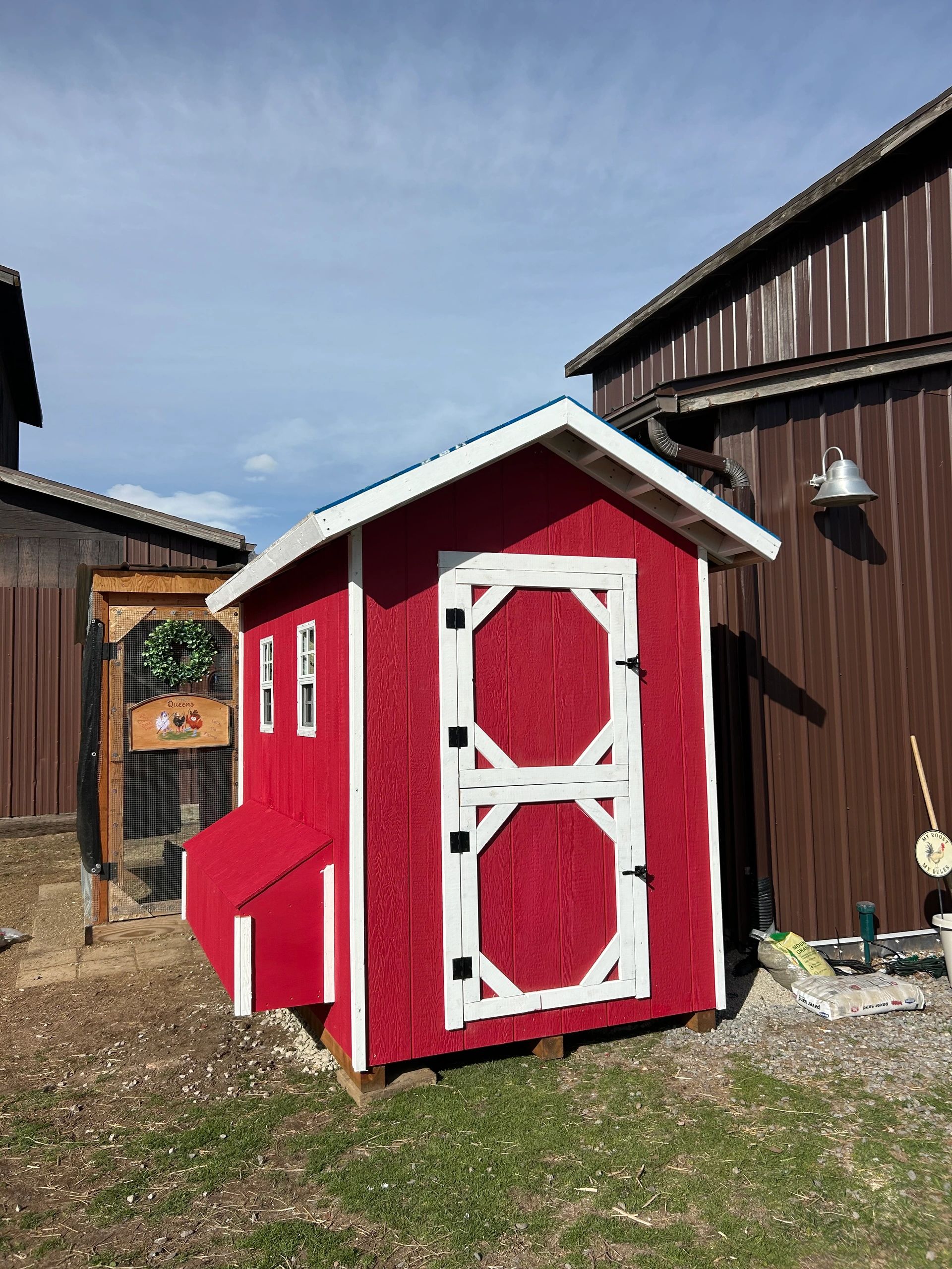 A bright red chicken coop with white trim outside a brown barn under a clear sky.