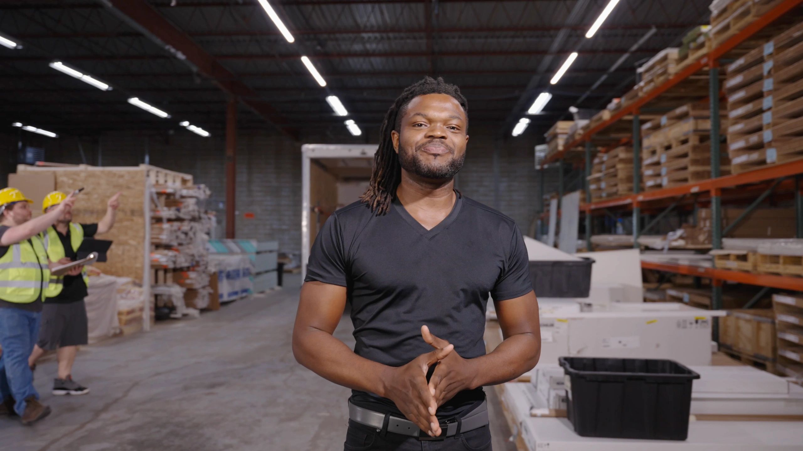 A man in a black shirt stands confidently in a warehouse with workers in safety gear in the background.