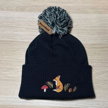 Black knit pom-pom beanie with fox and mushroom embroidery.