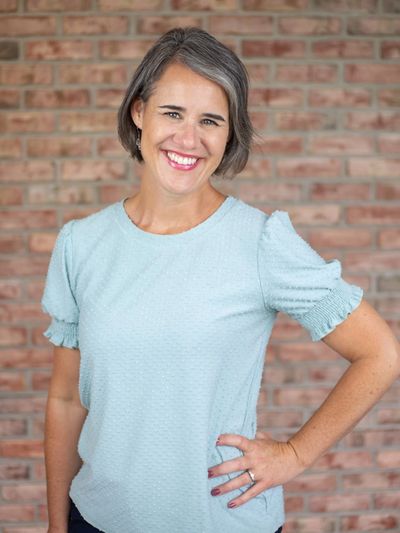 Smiling woman with gray hair in a light blue blouse stands against a brick wall.
