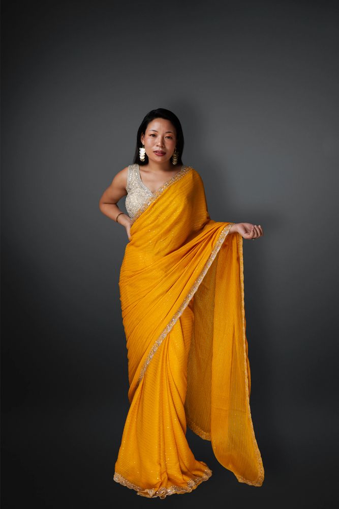 Woman posing in a vibrant yellow saree with a silver blouse against a gray backdrop.
