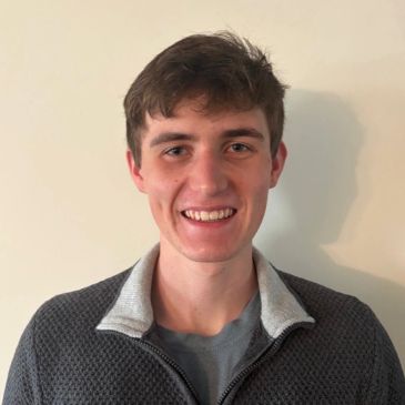 A young man with brown hair smiling against a plain wall.