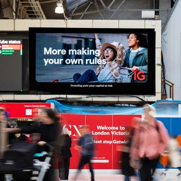 Busy commuters pass a vibrant London Victoria signage and a financial ad screen.