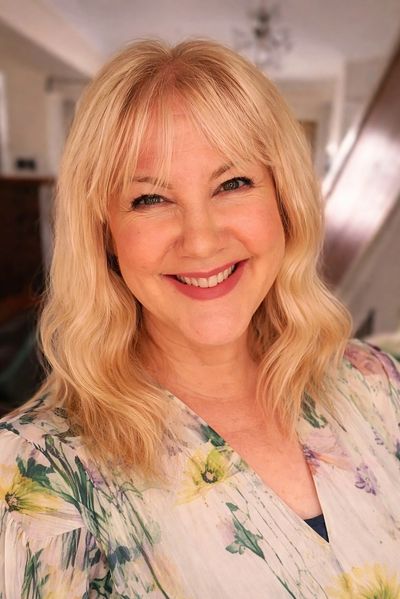 Smiling woman with blonde hair and floral blouse in a cozy home setting.