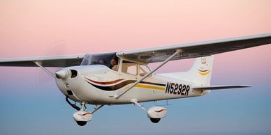 Small white airplane flying during sunset with two people inside.