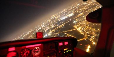 Night aerial view of a city from a cockpit with red instrument lights.